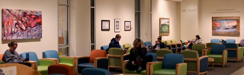 Several students studying in Art Alley with a variety of large, colorful paintings hanging on the wall.
