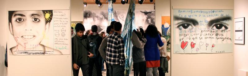 Several students listen to an artist describing their process. In the foreground are two large paintings depicting black and white close-ups of a child's face.