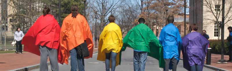 Seven people walking down a road in a line wearing capes of red, orange, yellow, green, blue, and purple fabric